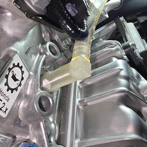 May include: Close-up of a silver engine component with a clear plastic tube and various bolts and fittings. The image shows a detailed view of the engine's internal parts, including a label with the text "Honda". The components are metallic and industrial in appearance.