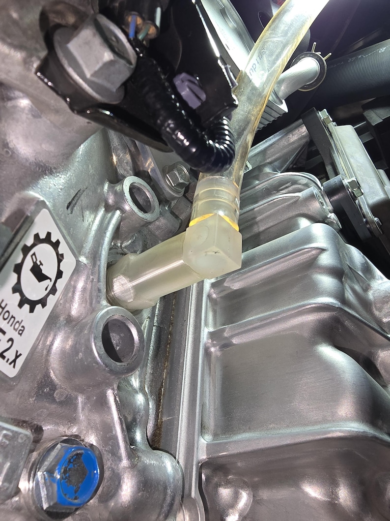 May include: Close-up of a silver engine component with a clear plastic tube and various bolts and fittings. The image shows a detailed view of the engine's internal parts, including a label with the text "Honda". The components are metallic and industrial in appearance.