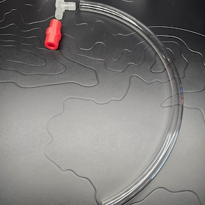 May include: A clear, flexible tube with a red connector and a white elbow connector. The tube is curved and appears to be made of plastic. The connectors are made of plastic. The background is a dark gray surface.