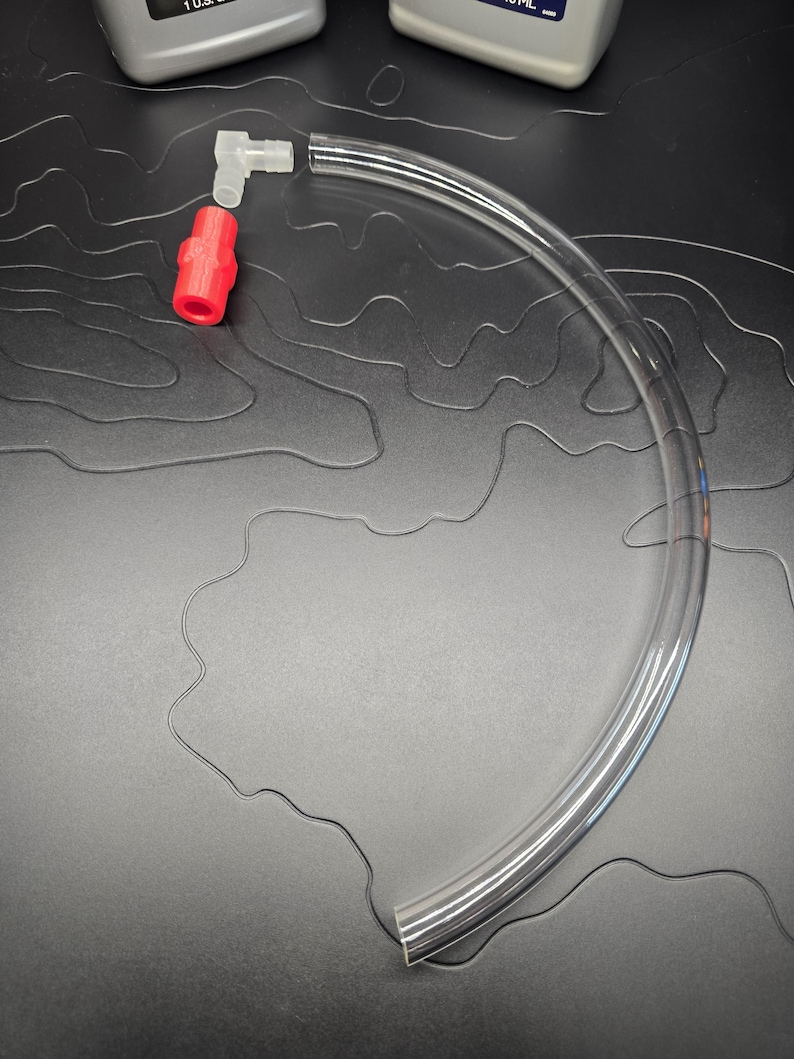 May include: A clear, flexible tube with a red connector and a white elbow connector. The tube is curved and appears to be made of plastic. The connectors are made of plastic. The background is a dark gray surface.