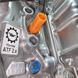 May include: Close-up of a car engine component with an orange cap and a blue bolt. The engine block is silver with the text "Honda ATF 2.x" and "ATF LEVEL" visible on a sticker and the engine block.