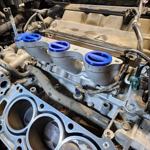 May include: Close-up of a car engine with a silver intake manifold and blue intake manifold covers. The engine is dirty and has a lot of wires and hoses.