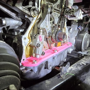 May include: Close-up of an engine's internal components, showcasing metallic parts, a timing chain, and a bright pink gasket. The image highlights the intricate details of the engine's mechanics, with bolts and other hardware visible.