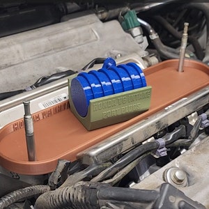 May include: A close-up of a Honda K20 intake port plugs kit. The kit includes a set of blue intake port plugs and a green plastic holder with the text "Honda K20 Intake Port Plugs". The kit is installed on a car engine.