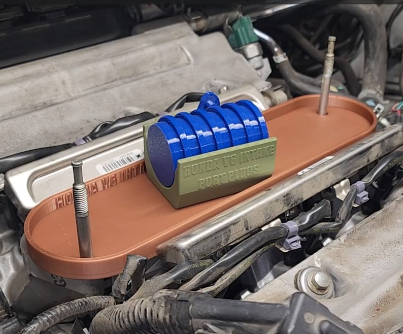 May include: A close-up of a Honda K20 intake port plugs kit. The kit includes a set of blue intake port plugs and a green plastic holder with the text "Honda K20 Intake Port Plugs". The kit is installed on a car engine.