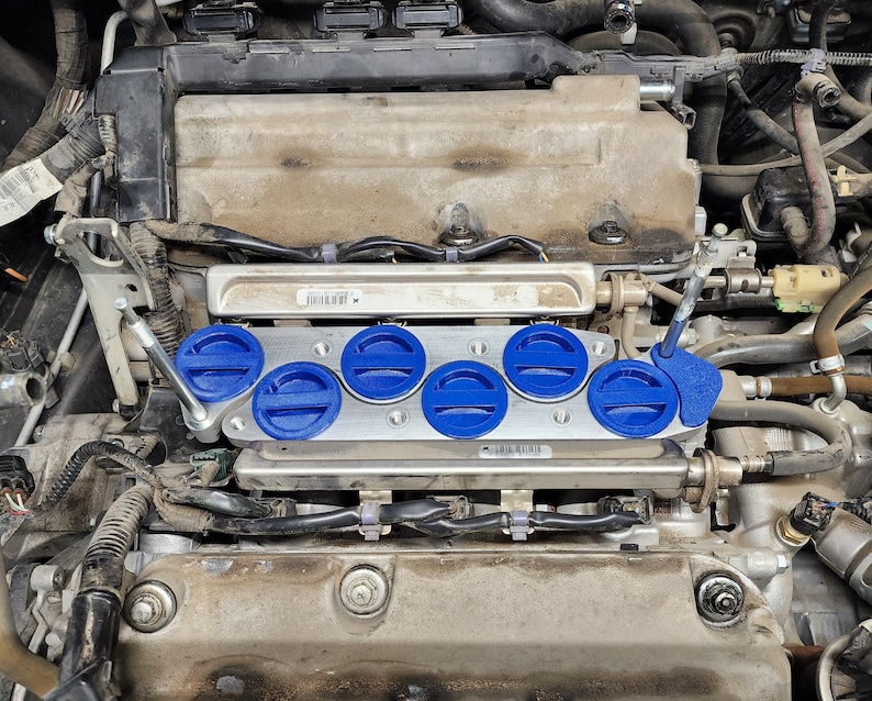 May include: Close-up of a car engine with a silver metal manifold and six blue fuel injectors. The injectors are connected to a black fuel rail.