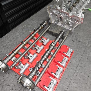 May include: A silver engine cylinder head positioned near two red plastic organizers holding metal camshafts and rocker arms. The red organizers feature text that reads L15 VTEC RACER HOLDER. The components are metallic, showing intricate mechanical parts.