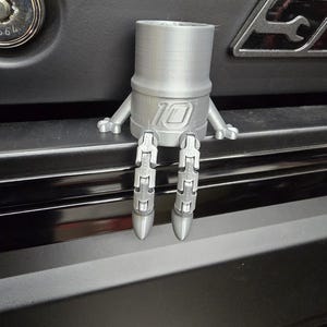 以下が含まれることがあります： 銀色の3Dプリントされたフィギュアで、本体に「10」の数字が入ったソケットレンチに似ています。このフィギュアは小さな腕と関節のある脚を持っており、風変わりな外観をしています。暗い表面に座っています。
