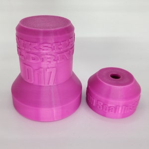Op de afbeelding: Twee roze 3D-geprinte plastic flessenstoppen. De grotere stop heeft de tekst "Oakshire Wild Drive 2017" aan de zijkant gedrukt. De kleinere stop heeft de tekst "Foam Seal Ins" aan de zijkant gedrukt.
