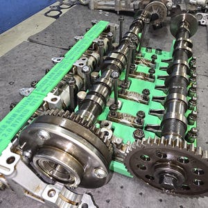 May include: A metallic engine camshaft assembly featuring two long, dark steel shafts with integrated gear sprockets, mounted on a bright green plastic organizer tray labeled "ROCKER ARM HOLDERS 2.0 HYBRID CYL 1" and "2.0 HYBRID CYL 2".
