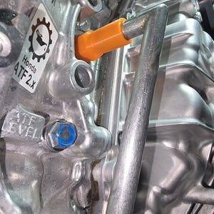 May include: Close-up of a silver engine component with an orange dipstick and a blue bolt. A sticker reads "Honda ATF 2.x" with a gear and oil can symbol. The text "ATF LEVEL" is also visible.