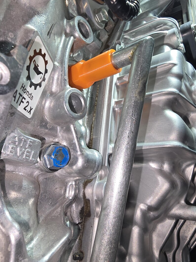 May include: Close-up of a silver engine component with an orange dipstick and a blue bolt. A sticker reads "Honda ATF 2.x" with a gear and oil can symbol. The text "ATF LEVEL" is also visible.