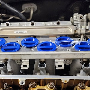 May include: Close-up of a car engine with blue valve covers. The engine is made of metal and has a silver finish. The valve covers are round and have a blue plastic cap.
