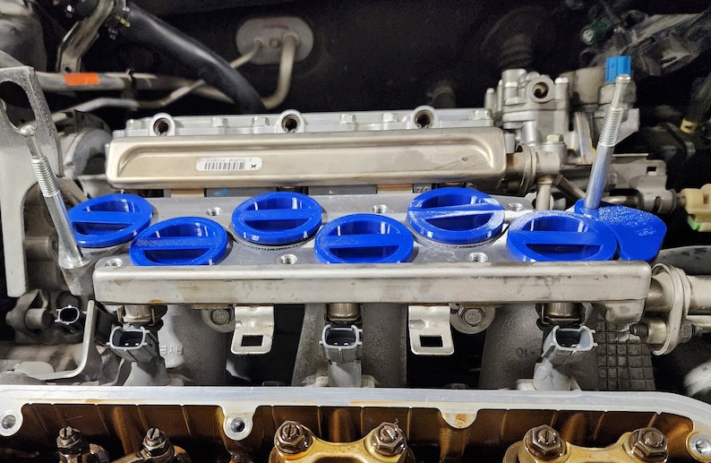 May include: Close-up of a car engine with blue valve covers. The engine is made of metal and has a silver finish. The valve covers are round and have a blue plastic cap.