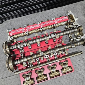 May include: A set of metallic engine components including two camshafts with gears, a long metal rod, and several rocker arm assemblies arranged on a bright red plastic organizer tray. The tray features red labels with white text reading K20 EARTH DREAMS ROCKER ARM HOLDERS.