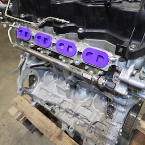 May include: A metallic automotive engine block featuring three distinct purple intake port gaskets installed along the side. The engine assembly includes a black plastic valve cover, various metal bolts, a fuel rail, and a silver metallic housing.
