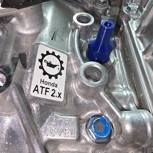 May include: Close-up of a silver engine component with a blue cap and a sticker that reads "Honda ATF 2.x". The sticker features a gear and oil can icon. The text "ATF LEVEL" is also visible.