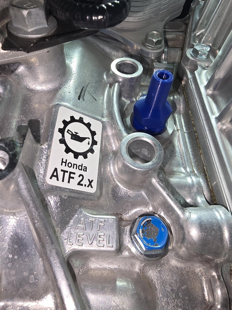 May include: Close-up of a silver engine component with a blue cap and a sticker that reads "Honda ATF 2.x". The sticker features a gear and oil can icon. The text "ATF LEVEL" is also visible.