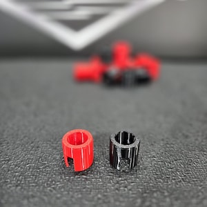 May include: Two small, cylindrical 3D-printed parts. One is bright red, the other is black. Both have a slot cut out of the side. The background shows more red parts, out of focus.