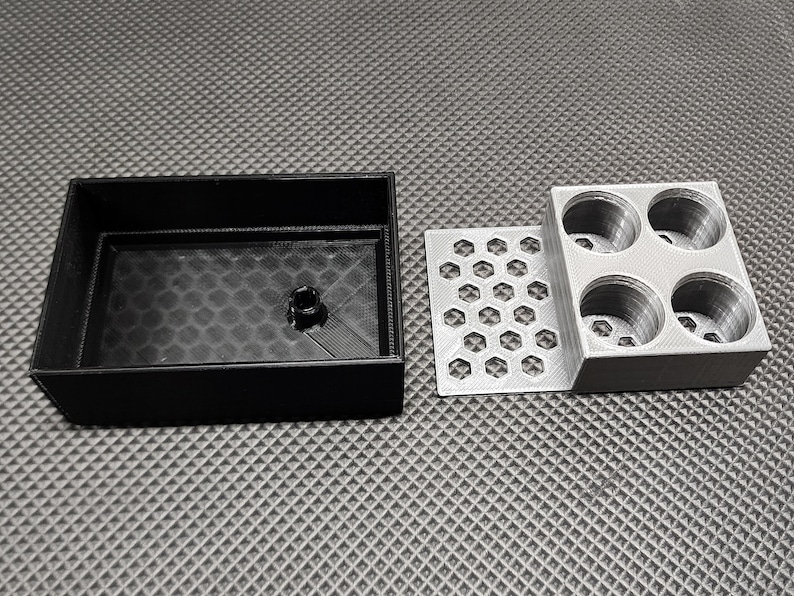 Peut inclure: Un conteneur rectangulaire noir avec un int&eacute;rieur textur&eacute;, une plaque argent&eacute;e en nid d'abeille et un bloc argent&eacute; avec quatre ouvertures circulaires. Les objets sont dispos&eacute;s sur une surface grise textur&eacute;e.