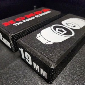 Puede incluir: Dos cajas de plástico negras con letras blancas y rojas. La caja superior tiene el texto "HONDA The Power of Dreams" y el número "22mm". La caja inferior tiene el número "19mm" y un gráfico blanco de una llave.