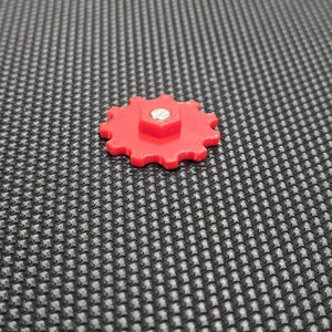 May include: A red plastic gear with a silver screw in the center. The gear has 12 teeth and is on a black textured surface.