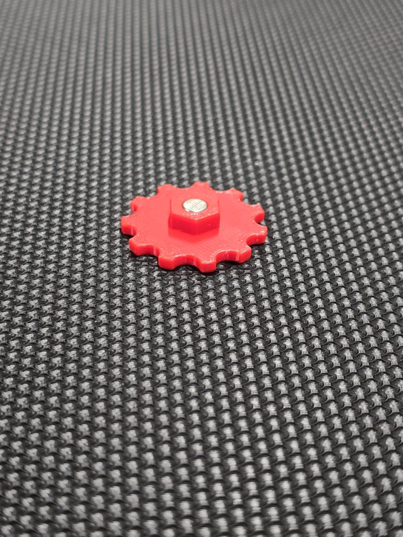 May include: A red plastic gear with a silver screw in the center. The gear has 12 teeth and is on a black textured surface.