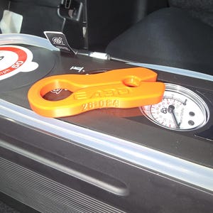 May include: A bright orange, flat, elongated plastic tool with a circular opening at one end and a slot at the other. The surface features embossed text reading EABD and 26-084. It is positioned on top of a black device with a circular pressure gauge.