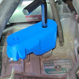 Peut inclure: Une pièce automobile en plastique bleu vif avec une clé hexagonale noire insérée. La pièce présente un design géométrique et est positionnée sur un composant moteur. Le logo Honda est visible en arrière-plan.