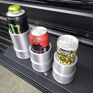 May include: Three aluminum drink holders with energy drink cans. The cans are black with green accents, red with white text, and yellow with black text. The holders are silver and cylindrical, designed to fit the cans snugly.