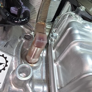 May include: Close-up of an engine component with a clear, angled tube connected to a metal housing. The tube has markings and is filled with a light-colored fluid. The engine parts are silver and have various bolts and fittings.