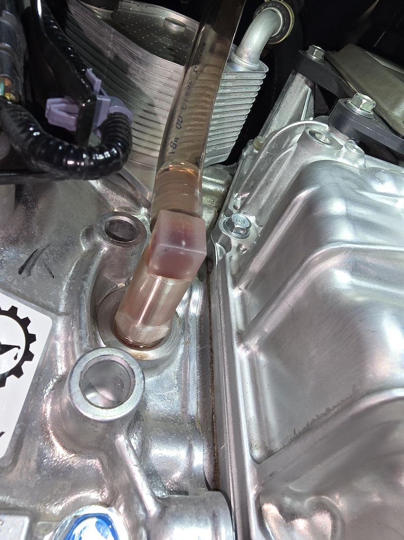 May include: Close-up of an engine component with a clear, angled tube connected to a metal housing. The tube has markings and is filled with a light-colored fluid. The engine parts are silver and have various bolts and fittings.