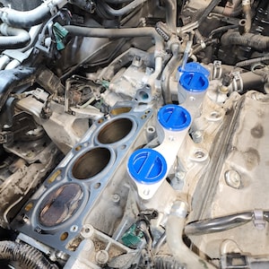 May include: Close-up of a car engine with a silver metal cylinder head and blue plastic valve covers. The cylinder head has three open cylinder bores and a gasket in place. The valve covers are labelled with the letter "E".