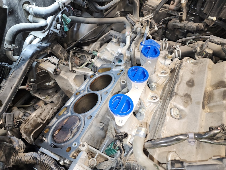 May include: Close-up of a car engine with a silver metal cylinder head and blue plastic valve covers. The cylinder head has three open cylinder bores and a gasket in place. The valve covers are labelled with the letter "E".