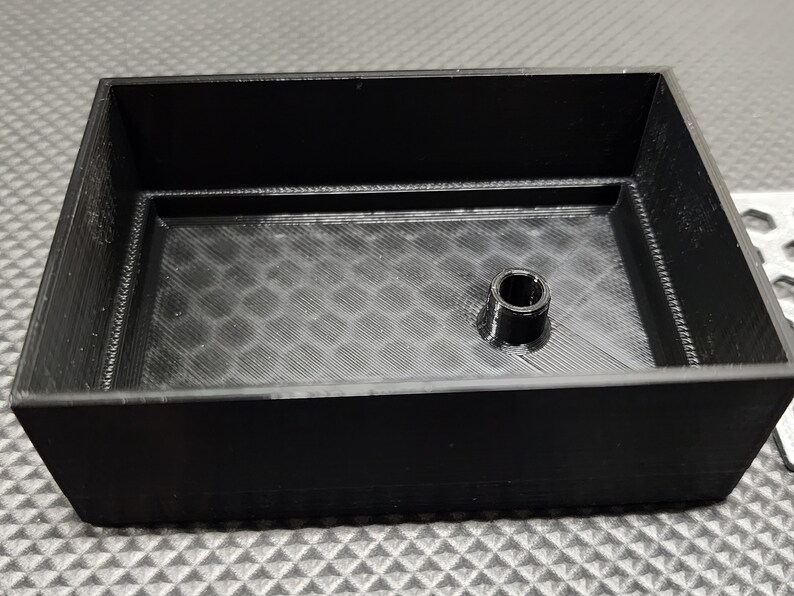 Peut inclure: Un r&eacute;cipient rectangulaire noir avec un cylindre central sur&eacute;lev&eacute;. L'int&eacute;rieur pr&eacute;sente un motif en nid d'abeille textur&eacute;. Les parois du r&eacute;cipient sont inclin&eacute;es, et le design est simple et fonctionnel.