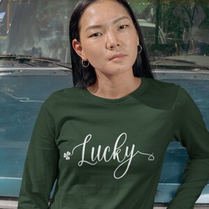 May include: Dark green long-sleeved top featuring the word "Lucky" in white script, with a shamrock and heart detail. The shirt is made of a soft material.