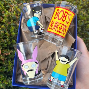 Może przedstawiać: Zestaw czterech przezroczystych kieliszk&oacute;w do w&oacute;dki z postaciami i logo serialu animowanego "Bob's Burgers". Każdy kieliszek ma inny wz&oacute;r postaci, w tym Tinę, Louise, Boba i logo serialu. Kieliszki znajdują się w niebieskim pudełku.