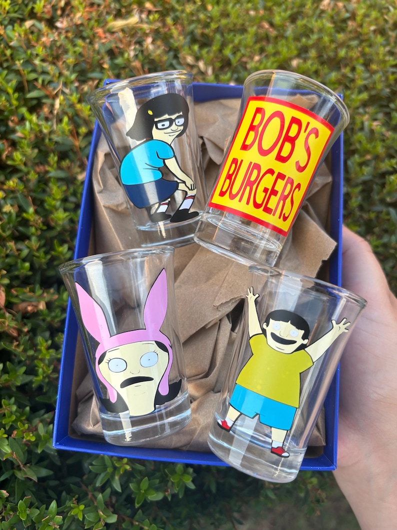 Może przedstawiać: Zestaw czterech przezroczystych kieliszk&oacute;w do w&oacute;dki z postaciami i logo serialu animowanego "Bob's Burgers". Każdy kieliszek ma inny wz&oacute;r postaci, w tym Tinę, Louise, Boba i logo serialu. Kieliszki znajdują się w niebieskim pudełku.