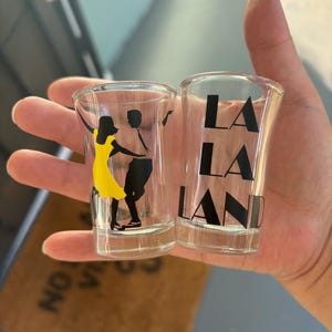 Peut inclure: Deux verres à shot transparents avec les mots "LA LA LAND" imprimés en noir. Un verre présente la silhouette d'un couple dansant, la femme portant une robe jaune. Les verres sont tenus dans une main.