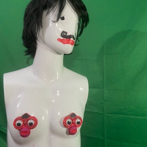 Puede incluir: Un maniquí blanco con cabello negro y un bigote y labios pintados. Dos juguetes de plástico rojo con ojos saltones y una boca roja están unidos al pecho del maniquí.