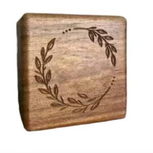 Puede incluir: Una caja de madera cuadrada con un dise&ntilde;o de corona decorativo grabado en la parte superior. La corona est&aacute; hecha de hojas y ramas.
