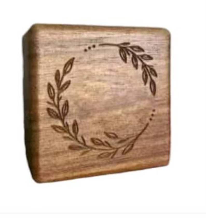 Puede incluir: Una caja de madera cuadrada con un dise&ntilde;o de corona decorativo grabado en la parte superior. La corona est&aacute; hecha de hojas y ramas.