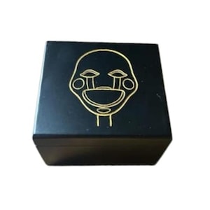 May include: A black square box with a gold outline of a stylized face on the top. The face has large eyes, a wide mouth, and tear-shaped markings. The box is likely made of wood or a similar material.