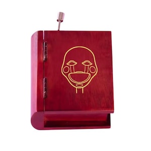 May include: A burgundy-colored wooden box with a hinged lid and a silver metal pull. The front of the box features a gold outline of a puppet face. The box is designed to look like a book.