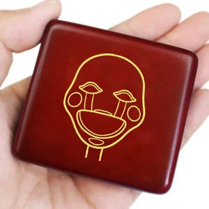 May include: A square, wooden box with a deep red finish. The box features a gold outline of a stylized face with large eyes and a wide smile. The box is held in a person's hand.