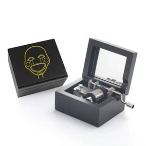 以下が含まれることがあります： 黒いオルゴール。ヒンジ付きの蓋と透明な窓が付いています。箱は開いており、内部のメカニズムが明らかになっています。黄色の顔のデザインが施された別の黒い箱が隣にあります。