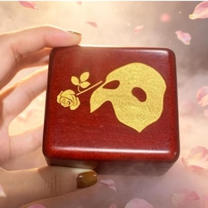 May include: A square, wooden box with a dark red finish. The box features a gold-colored mask and rose design on the top. The box is held by a hand, with a soft, blurred background of pink and white petals.