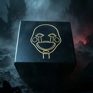 Puede incluir: Una caja cuadrada negra que presenta una ilustración de líneas doradas de un rostro de marioneta estilizado en la superficie superior. El diseño del rostro incluye mejillas circulares, ojos almendrados con marcas de lágrimas y una boca ancha y curva.
