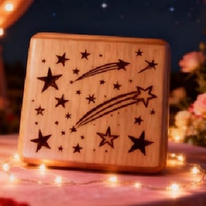 May include: A wooden music box with a star design. The box is light brown with etched black stars and shooting stars. The box is square and sits on a pink surface with string lights. The background includes flowers and a dark blue sky.