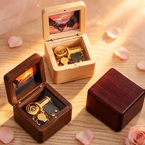 May include: Three square wooden music boxes. Two are open, revealing gold-toned mechanical movements and small photographs inside the lids. One box is made of light wood, the other of dark wood. A third dark wooden box is closed and sits to the right.